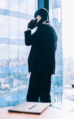 Takım elbiseli şık siyahi bir adamın modern ofisin penceresinin yanında çeşitli meslektaşlarıyla masa başında telefon görüşmesi yaparken yan görüntüsü.