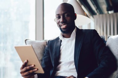 Yakışıklı, takım elbiseli, elinde tablet, koltukta oturmuş ofisin koridorundaki kameraya gülümsüyor.