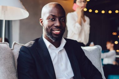 Koltukta otururken tablet tutan ve ofis koridorundaki kameraya güvenle bakan takım elbiseli, zarif, Afro-Amerikan bir adam.