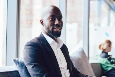 Ofis koridorundaki koltukta oturan şık, yakışıklı, Afro-Amerikan bir adam dizüstü bilgisayarla kameraya gülümsüyor.