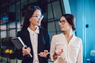 Zarif giysiler içinde çok kültürlü güzel genç kadınlar ofis koridorunda birbirleriyle konuşurken ellerinde dosyalar ve telefonlarla yürüyorlar.