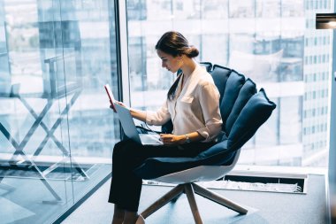 Modern ofis dizüstü bilgisayarında koltukta oturan ve çalışırken cep telefonu kullanan güzel bir kadının yan görüntüsü.