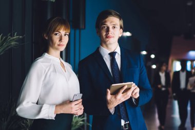 Akıllı telefonlu ciddi bir iş kadını ve tabletli zarif genç bir adam ofis koridorunda birlikte kameraya özgüvenle bakıyor.