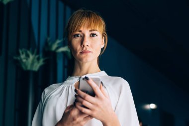 Zarif takım elbiseli, kendine güvenen güzel bir kadının elinde cep telefonuyla ofiste duran kameraya ciddi ciddi bakmasından.