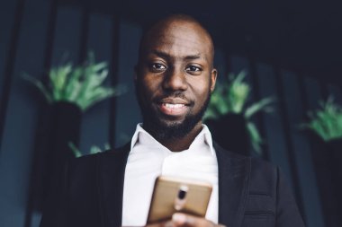 Takım elbiseli, kendine güvenen Afrikalı Amerikalı bir adamın elinde cep telefonuyla ofis koridorundaki kameraya bakmasından.