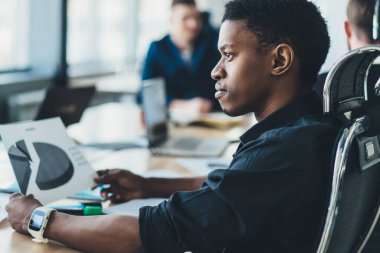 Düşünceli Afrikalı Amerikalı ofis çalışanı arka planda meslektaşlarıyla toplantı masasında oturuyor.