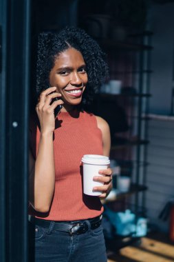 Yüzünde samimi bir gülümseme ile kamera bakıyor ve en iyi arkadaşı ile akıllı telefon uygulaması ile iletişim keyfini el sıcak içki ile neşeli milenyal hipster kız yarı uzunlukta portre