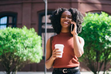 Güneşli yaz gününde açık havada poz kafein içecek ile mutlu Afrikalı Amerikan hipster kız yarı uzunlukta portre, Genç kadın online uygulama üzerinden Smartphone konuşma zevk