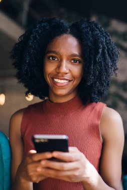 Cep telefonu, mutlu milenyal kadın tutan akıllı telefon aracı ile online iletişim sırasında arkadaşınızın SMS cevap beklerken kamerada gülümseyen neşeli koyu tenli kadın portre yakın