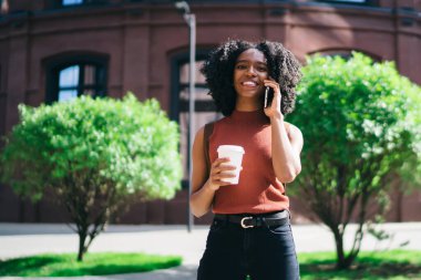 Güneşli yaz gününde açık havada poz kafein içecek ile mutlu Afrikalı Amerikan hipster kız yarı uzunlukta portre, Genç kadın online uygulama üzerinden Smartphone konuşma zevk