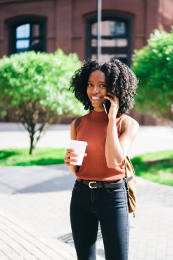 Karanlık tenli hipster öğrenci kahve molası sırasında akıllı telefon konuşma zevk açık havada, cep telefonunda uygulama üzerinden konuşurken rahat giyim kamera gülümseyen giyinmiş mutlu kadın