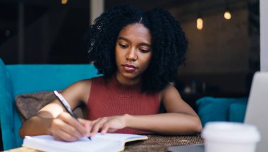 Esmer tenli hippi kız, araştırma ve öğrenim için modern teknolojiyle masaya otururken bilgi notunda yazıyor.