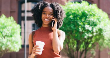 Gündelik giyinmiş, dışarıda boş zamanlarında kameraya gülümseyen neşeli yüzlü bir genç kızın yarım boy portresi. Gezintide 4G 'ye bağlı mutlu siyah kadın arkadaşını arıyor.