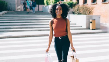 Başarılı esmer kadın portresi. Günlük kıyafetlerle süslenmiş. Alışverişten heyecanlanmış. Elinde kağıt torbalarla sokakta yürüyen pozitif bir kadın.