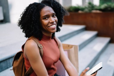 Modern dijital cihazları olan mutlu bir genç kızın portresi takipçilerle çevrimiçi iletişim kurarken gülüyor, neşeli esmer tenli kadın şehir ortamında dinleniyor.