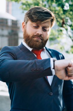 İş merkezine karşı caddede duran kırmızı kravatlı ciddi sakallı iş adamı öğle arasında kol saatine bakıyor.