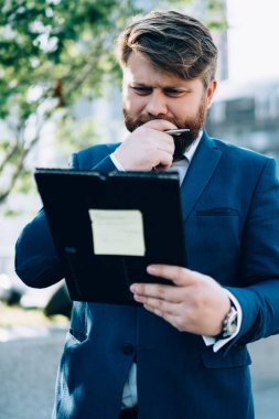 Sakallı, takım elbiseli düşünceli bir işadamı iş merkezinin caddesinde duruyor. Elinde klasör okuyup, çenesini çıkmaza sokuyor.