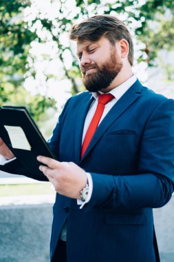 Ciddi sakallı iş adamı takım elbise giymiş ofis binalarının yanında duruyor. Yemek molasında toplantı için hazırlanan klasördeki belgeleri okuyor.
