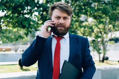 Endişeli sakallı, şık takım elbiseli bir işadamı iş merkezinin yanında elinde dosyayla kameraya bakarak telefonda konuşuyor.