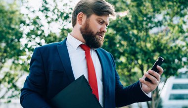 Stresli, sakallı, takım elbiseli iş adamı cep telefonuna bakıyor, mesaj okuyor ve iş merkezinin yakınındaki bir sokakta elinde klasörle mesajlaşıyor.