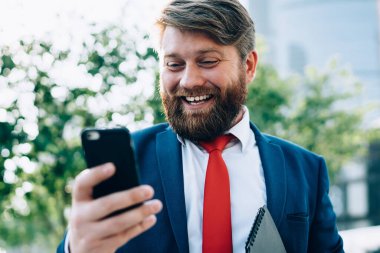 Zarif takım elbiseli, kırmızı kravatlı, akıllı telefon kullanarak partneriyle sohbet eden, modern şehrin parkında yürüyen neşeli, güler yüzlü adam.