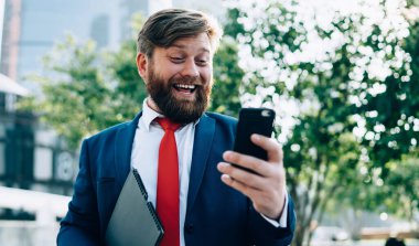 Zarif elbiseli, kırmızı kravatlı mutlu adam modern şehir parkında takılan iş arkadaşıyla konuşurken eğleniyor.