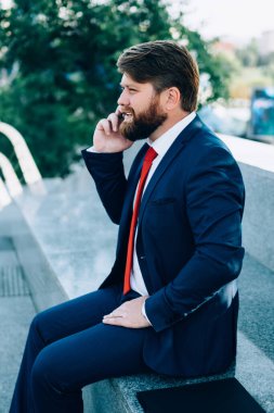Şık takım elbiseli, şık ve kırmızı kravatlı, modern şehrin parkında çitin üzerinde oturan akıllı telefon kullanarak iş arkadaşlarıyla iş problemlerini tartışan stresli bir erkek.