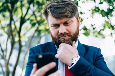 Takım elbiseli, kırmızı kravatlı ve çenesini düşüncelerle ovuşturan patron şehir parkında çitin üzerinde oturan bir projede cep telefonu kullanıyor.