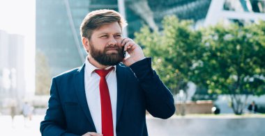 Koyu renk şık takım elbiseli, kırmızı kravatlı düşünceli sakallı adam iş merkezinin meydanında cep telefonuyla konuşurken dikkatle dinliyor.
