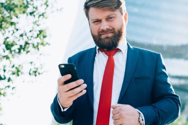 Koyu renk takım elbiseli, kırmızı kravatlı, elinde cep telefonu olan dalgın bir iş adamı iş merkezindeki kameraya dikkatle bakıyor.