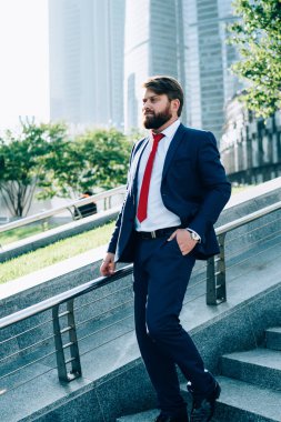 Sakallı, düşünceli, zarif takım elbiseli ve kırmızı kravatlı işadamının yan görünüşü. Merdivenlerde el ele yürüyorlar. Arka planda cam bina koluna yaslanıyorlar.