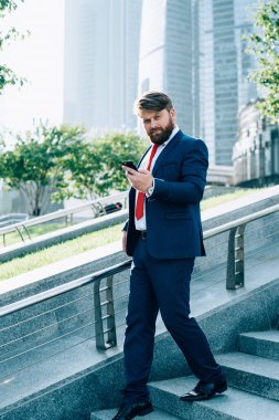 Sakallı, düşünceli, şık takım elbiseli, kırmızı kravatlı bir işadamı elinde cep telefonuyla merdivenlerden inip kameraya bakarken.