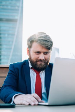Modern şehrin sokağındaki ahşap masada otururken dizüstü bilgisayarını kullanarak resmi kıyafet giyen yönetici odaklı sakallı adam.
