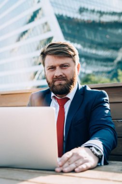 Kırmızı kravatlı, kaşlarını çatmış bir adam iş için dizüstü bilgisayar kullanıyor. İş yerindeki masada oturan kameraya bakıyor.