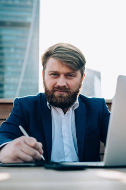 Resmi olarak yönetici odaklı sakallı adam dizüstü bilgisayarla uzaktan çalışma fikirleri yazıp modern şehrin parkındaki masada oturuyor.