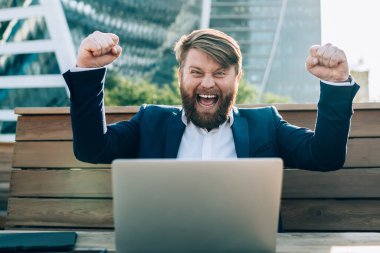 Resmi giysiler içinde ellerini kaldırmış neşeli başarılı patron kameraya bakıyor iş bölgesinde masada oturarak kazanmanın keyfini çıkarıyor.