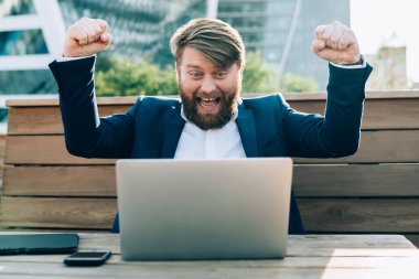 Mavi takım elbiseli neşeli ve enerjik. Kollarını kaldırmış, dizüstü bilgisayarla şehirde açık terasta oturuyor.