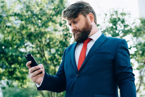 Takım elbiseli ve kırmızı kravatlı konsantre sakallı işadamı iş merkezinin yanındaki klasörde duran akıllı telefondan mesaj okuyor.