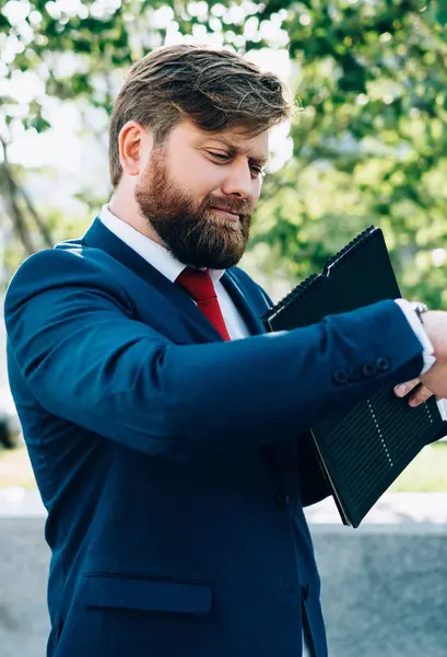 Stresli, takım elbiseli, ciddi sakallı iş adamı ofis binasının önünde duruyor. Kol saatine bakıyor ve önemli bir müşteri bekliyor.
