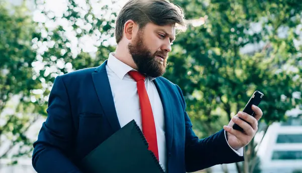 Stresli, sakallı, takım elbiseli iş adamı cep telefonuna bakıyor, mesaj okuyor ve iş merkezinin yakınındaki bir sokakta elinde klasörle mesajlaşıyor.