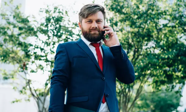 Takım elbiseli ve kırmızı kravatlı ciddi bir iş adamı ofis binasının önünde dikilip kameraya bakarken telefonla konuşuyor.