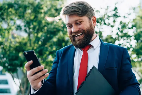 Kırmızı kravatlı neşeli sakallı erkek, modern şehrin parkında telefonla partnerini arıyor.