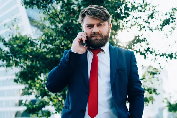 Koyu renk takım elbiseli, kırmızı kravatlı neşeli bir adam cep telefonuyla konuşurken ve iş merkezinin karesinde el ele kameraya bakarken dinliyor.