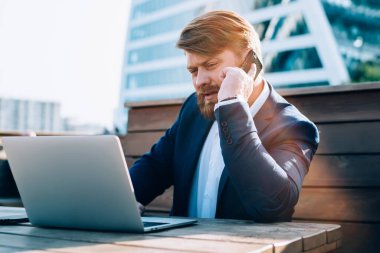 Güneş ışığı altında konsantre sakallı, ciddi bir erkek. Laptop seyrediyor. Akıllı telefondan konuşuyor.