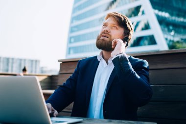 Düşünceli, kafası karışmış resmi giyinmiş, akıllı telefon sörf laptopuyla şehir merkezindeki ahşap masada oturan aynalı gökdelenlerle konuşan bir erkek.