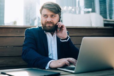 Mavi ceketli ve beyaz gömlekli sakallı adam şehir merkezindeki kafedeki ahşap masada oturmuş cep telefonu sörf laptopuyla konuşuyor.