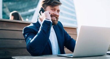 Takım elbiseli memnun bir adam telefonda konuşuyor ve kahkahalarla gülüyor bir yandan da sokak kafesinde kameraya bakıyor.