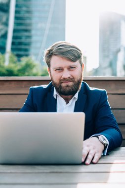 Şehir binalarının arka planında sokak kafesinde otururken dizüstü bilgisayarla çalışan yakışıklı şık bir adam.