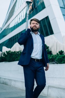 Takım elbiseli ciddi bir yönetici sokakta yürüyor ve telefonla konuşuyor cam binanın merkezine bakıyor.