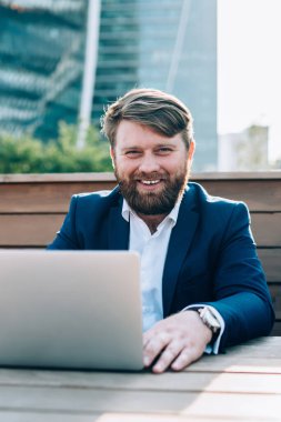 Sakallı, mutlu ve kendinden emin bir adam kameraya gülümsüyor. Şehir arka planında, sokak kafesinde bilgisayarla çalışıyor.